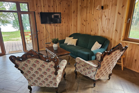 Lodge living room with wooden walls, green sofa, patterned armchairs, wall-mounted TV, and garden view.