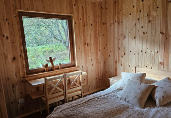 Dormitorio en una cabaña de madera con gran ventana, cama cómoda y dos sillas junto a un escritorio.