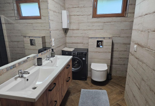 Modern badkamer in een lodge met dubbele wastafel, wasmachine onder plank, toilet en grote ramen.