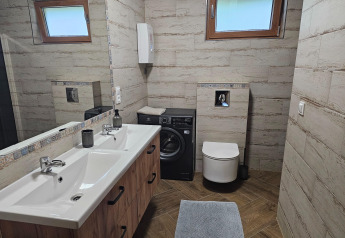 Moderne lodgebadkamer met dubbele lavabo, wasmachine onder een plank, wc en twee grote ramen.