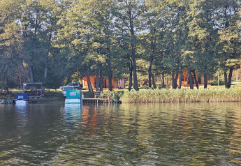 Imagen de un albergue en la orilla del lago, rodeado de árboles y barcos atracados bajo el sol.