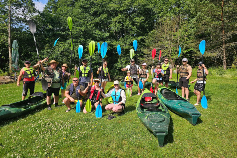 Group of people with paddles and life vests, ready to kayak near a lodge in a lush, green setting.