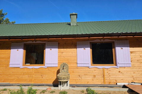 Vakantiehuisje met groen dak, lavendelkleurige luiken en buitenfontein onder blauwe lucht in Grąbczyn, Denemarken.