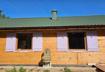 Cabaña vacacional con techo verde, contraventanas lilas y fuente exterior bajo cielo azul en Grąbczyn, Dinamarca.