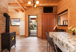 Træinteriør af hyggelig lodge med brændeovn, spisebord, skab og udsigt til udendørs område.