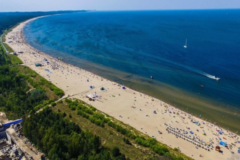 Vista aérea de una larga playa de arena con aguas azules, bosque verde y muchas personas disfrutando.