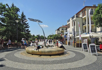 Straßenszene bei Ferienhäusern in Swinoujście, Menschen entspannen an moderner Springbrunnen-Skulptur.
