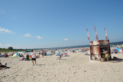 Playa concurrida con personas, tiendas de colores y una pequeña cafetería bajo un cielo azul claro.