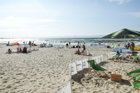 Folk nyder solen på en sandstrand ved havet, med liggestole, parasoller og vindskærme omkring.