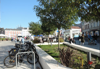 Escena urbana en Swinoujście con bicicletas, árboles y gente paseando y conversando en un día soleado.