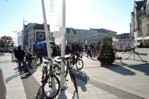 Plaza concurrida en Świnoujście con bicicletas, personas, carteles electorales y edificios al fondo.