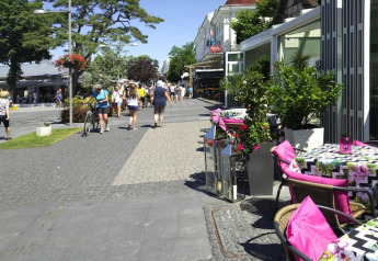 Cafetería exterior con cojines coloridos y flores; gente paseando por calle peatonal en Rewal, Alemania.
