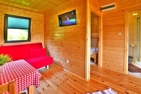 Interior de una cabaña de madera en Mielno con sofá rojo, mesa de comedor y vista al baño.