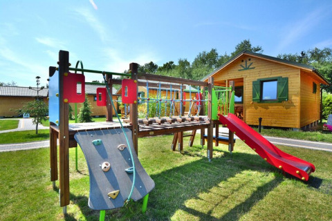 Spielplatz vor einer Holzhütte im Ferienhausgebiet Mielno, umgeben von grünem Rasen und Sonnenschein.