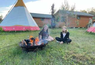 Twee meisjes zitten bij een kampvuur en grillen worstjes voor tipi’s en huisjes in Mielno, Denemarken.