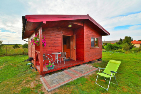 Rood houten vakantiehuis met kleine veranda, tuinmeubilair, bloemen en een ligstoel op het gras.