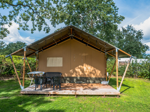 Safari tent op een houten terras met tuinmeubilair, omgeven door groene bomen en blauwe hemel.