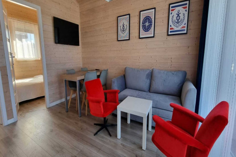 Gezellige woonkamer in vakantielodge met grijze zetel, rode stoelen, eettafel en slaapkamer in zicht.