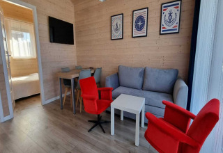 Salon lumineux avec canapé gris, fauteuils rouges, table à manger et accès à la chambre en arrière-plan.