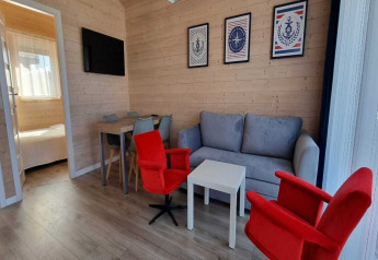 Helles Wohnzimmer in einem Ferienhaus mit grauem Sofa, roten Sesseln und Zugang zum Schlafzimmer.