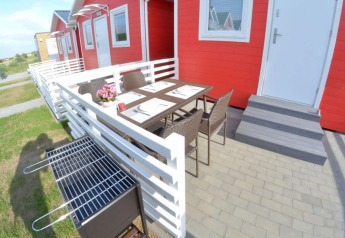 Udendørs terrasse med spisebord, stole og grill foran røde enkeltplans feriehytter i Sarbinowo, Danmark.