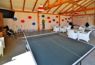 Overdekte recreatieruimte met tafeltennistafel, witte stoelen, tafels en kleurrijke wandversieringen.
