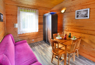 Gezellige blokhut met paarse zetel, houten eettafel, stoelen en een helder raam met vitrage.