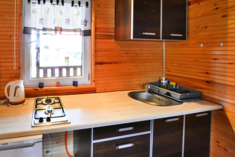 Kleine keukenhoek in houten chalet met gasvuur, spoelbak, waterkoker en raam met wit gordijn.