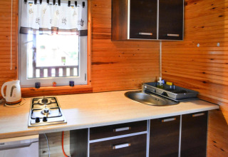 Kleine keukenhoek in houten chalet met gasvuur, spoelbak, waterkoker en raam met wit gordijn.