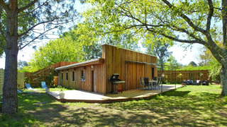 Ferienhaus Le Porge mit Holzfassade, Terrasse, Grill und Gartenmöbeln in grünem Garten.