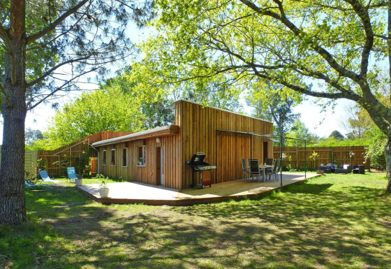 Maison de vacances Le Porge, lodge en bois avec terrasse, barbecue et mobilier de jardin sous les arbres.