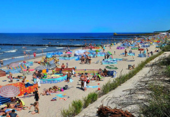 Strandbillede fra Ustronie Morskie med mange mennesker, farverige parasoller og badegæster ved havet.