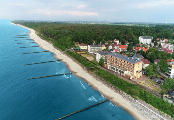 Luftfoto af en strand og kystby ved Ustronie Morskie, med huse, hoteller og blå hav ved skovgrænsen.