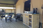 Veranda esterna con area salotto e mobile cucina davanti a una tenda safari di lusso attrezzata.