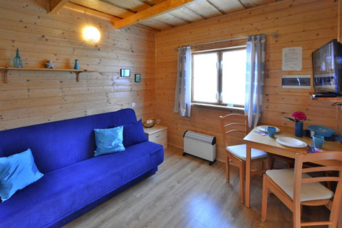 Cozy wooden cabin living area with blue sofa, dining table, window and TV at Holiday cottages Sarbinowo, Denmark.