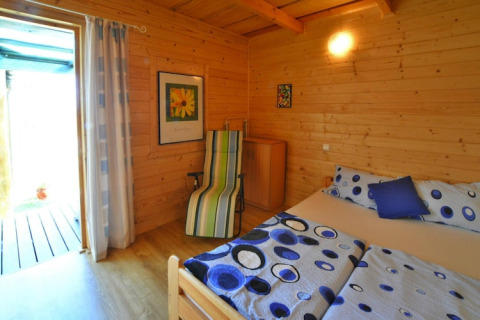 Gezellige houten slaapkamer met tweepersoonsbed, kleurrijk beddengoed, ligstoel en open deur naar buiten.