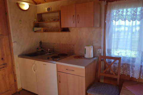 Kleine kitchenette in een houten huisje met keukenkastjes, waterkoker, gootsteen en kanten gordijn.