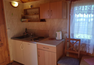 Kleine kitchenette in een houten huisje met keukenkastjes, waterkoker, gootsteen en kanten gordijn.
