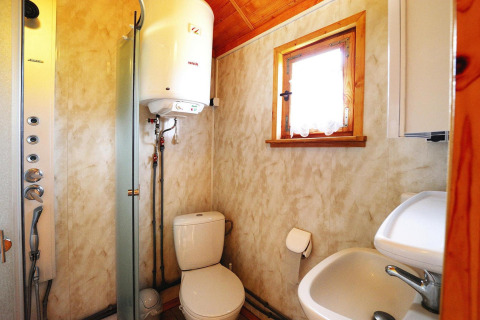 Compact cabin bathroom featuring a toilet, sink, water heater, shower and a small window with curtain.