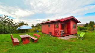 Holiday cottage Rusinowo with red wooden cabin, garden furniture and umbrella on a grassy lawn.