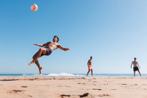 Three men play beach soccer by the sea, one performing a bicycle kick under a clear blue summer sky.