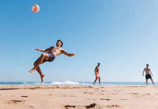 Three men play beach soccer by the sea, one performing a bicycle kick under a clear blue summer sky.