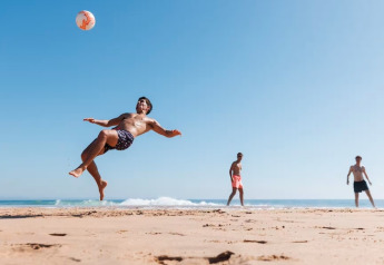Tres hombres juegan fútbol playa junto al mar, uno realiza una chilena bajo un cielo azul despejado.