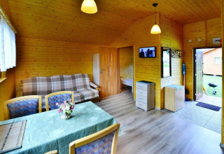 Interior view of a cozy wooden cabin with dining table, plaid sofa, open bedroom, and wall-mounted TV.