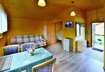 Interieur van een gezellige houten chalet met eettafel, zetel, open slaapkamer en televisie aan de muur.