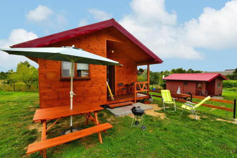 Wooden cabin with red roof, picnic table, umbrella, and grill on grassy lawn, sunny peaceful atmosphere.
