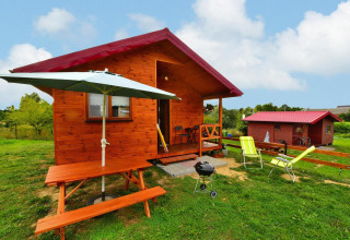 Wooden cabin with red roof, picnic table, umbrella, and grill on grassy lawn, sunny peaceful atmosphere.