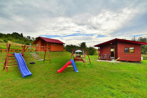 Playground with slides and swings in front of two wooden cabins on a grassy field under a cloudy sky.