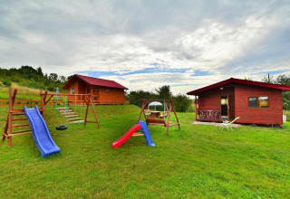Playground with slides and swings in front of two wooden cabins on a grassy field under a cloudy sky.