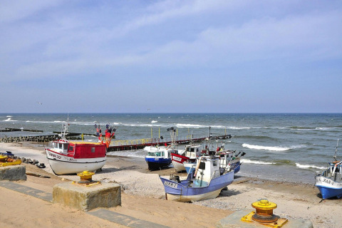 Fishing boats rest on a sandy shore by the sea with mild waves and a dock on a bright, sunny day.